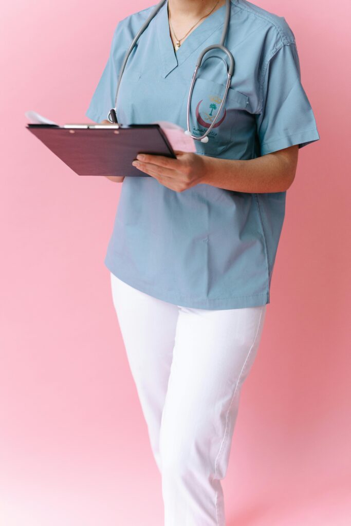 pexels-photo-7659867-7659867 Nurse in blue scrubs with a stethoscope holding a clipboard against a pink backdrop.